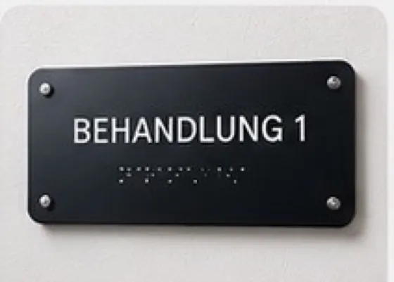 Schwarzes Türschild mit weißer Beschriftung Behandlung 1 und Braille-Zeile